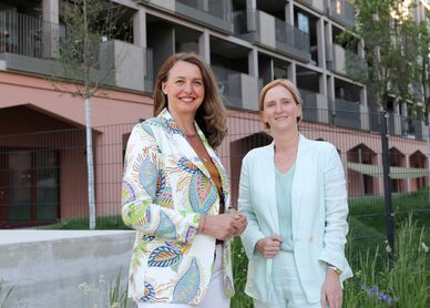 Zwei Frauen stehen vor einem modernen Wohngebäude, eine trägt einen gemusterten Blazer, die andere einen hellen Anzug