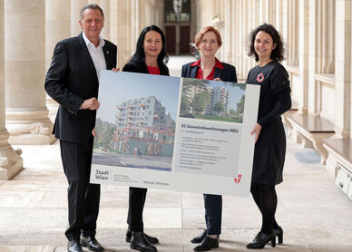 Drei weibliche und eine männliche Person stehen in den Arkaden des Wiener Rathauses und halten gemeinsam ein Schild auf dem Visualisierungen des Gemeindebau NEU zu sehen sind, sowie ein wenig Text