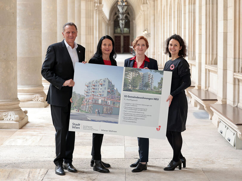 Drei weibliche und eine männliche Person stehen in den Arkaden des Wiener Rathauses und halten gemeinsam ein Schild auf dem Visualisierungen des Gemeindebau NEU zu sehen sind, sowie ein wenig Text