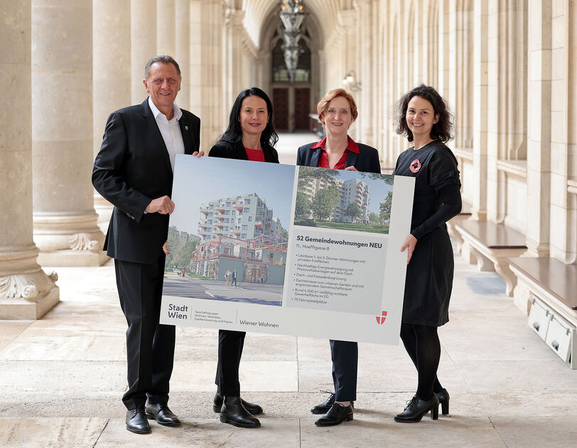 Drei weibliche und eine männliche Person stehen in den Arkaden des Wiener Rathauses und halten gemeinsam ein Schild auf dem Visualisierungen des Gemeindebau NEU zu sehen sind, sowie ein wenig Text