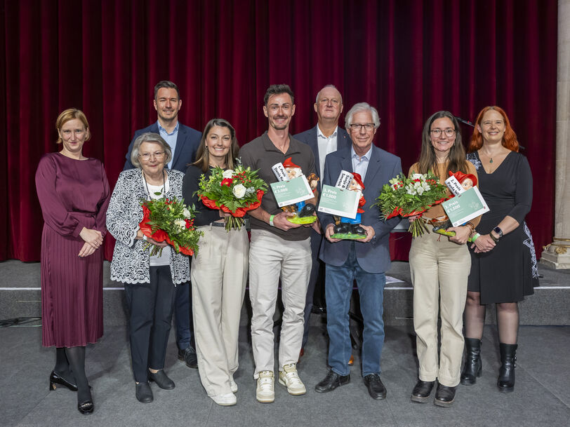 Gruppe von acht Personen vor rotem Vorhang und österreichischer Flagge, drei Personen halten Blumensträuße und Urkunden