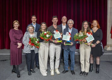 Gruppe von acht Personen vor rotem Vorhang und österreichischer Flagge, drei Personen halten Blumensträuße und Urkunden