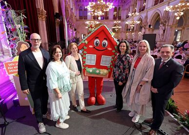Gruppe von neun Personen steht in festlich beleuchtetem Saal neben wienwohntbesser-Maskottchen, ein rotes Haus mit Gesicht und Blumenschmuck, sie halten ein Schild mit Text 'Wir sorgen für ein gutes Miteinander'