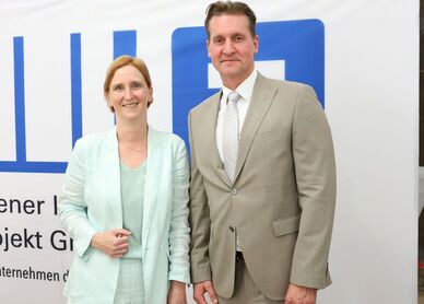 Zwei Personen in Anzügen stehen vor einem weißen Banner mit blauem Logo