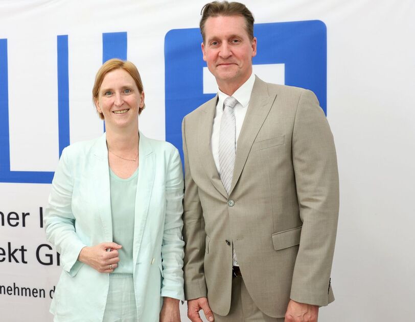 Zwei Personen in Anzügen stehen vor einem weißen Banner mit blauem Logo