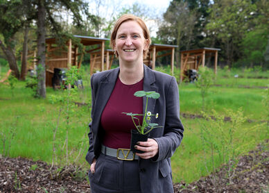 Person mit kurzem Haar und grauem Anzug hält einen kleinen Blumentopf mit einer Pflanze in einem Garten