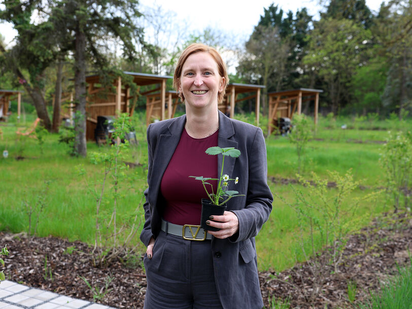 Person mit kurzem Haar und grauem Anzug hält einen kleinen Blumentopf mit einer Pflanze in einem Garten