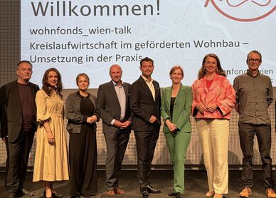 Gruppe von zehn Personen steht vor einer Präsentationswand mit dem Text 'Willkommen! wohnfonds_wien-talk Kreislaufwirtschaft im geförderten Wohnbau – Umsetzung in der Praxis'