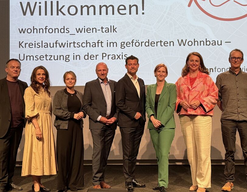 Gruppe von zehn Personen steht vor einer Präsentationswand mit dem Text 'Willkommen! wohnfonds_wien-talk Kreislaufwirtschaft im geförderten Wohnbau – Umsetzung in der Praxis'