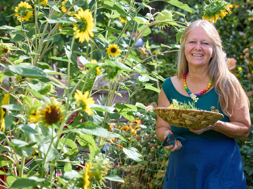 Frau mit langem Haar und bunter Halskette hält einen Korb mit Kräutern in einem Garten voller Sonnenblumen