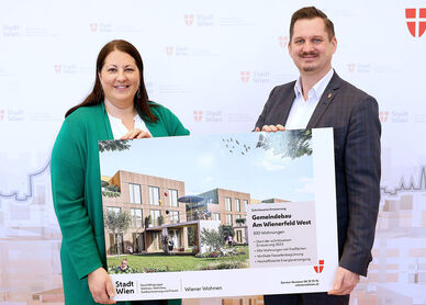 Eine Frau und ein Mann halten ein Schild mit der Visualisierung des neuen Gemeindebaus NEU in der Siedlung Wienerfeld West, erkennbar an der Beschriftung des Schildes. Sie lächeln in die Kamera.