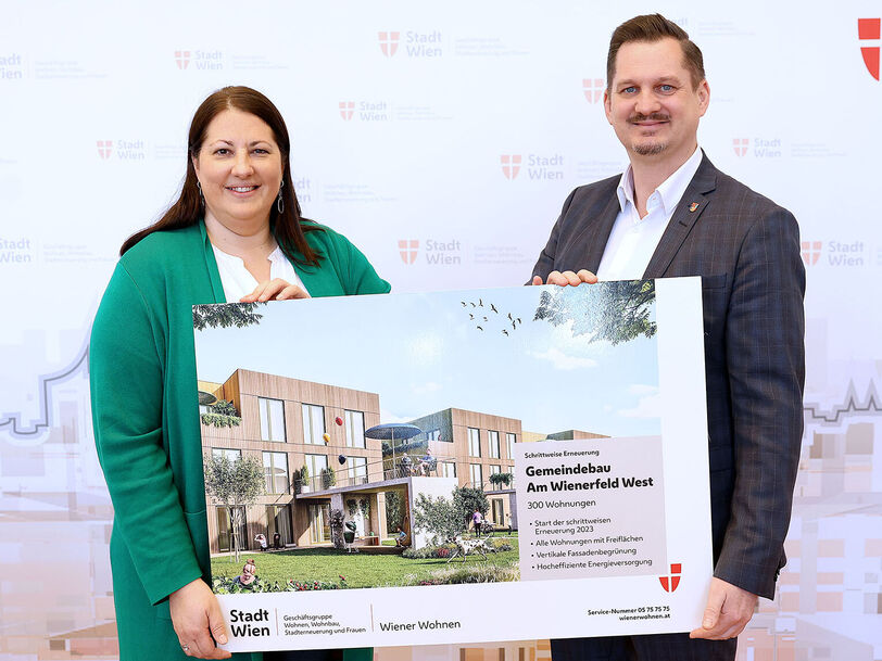 Eine Frau und ein Mann halten ein Schild mit der Visualisierung des neuen Gemeindebaus NEU in der Siedlung Wienerfeld West, erkennbar an der Beschriftung des Schildes. Sie lächeln in die Kamera.