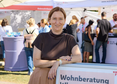 Frau lehnt an einem Schild mit der Aufschrift 'Wohnberatung Wien' auf einer Freiluftveranstaltung mit mehreren Menschen und Informationsständen im Hintergrund