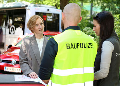 Drei Personen im Gespräch an einem Stehtisch, eine Frau trägt Blazer, ein Mann eine gelbe Weste mit Aufschrift "BAUPOLIZEI", eine weitere Frau trägt schwarze Weste