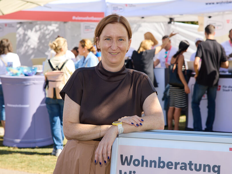Frau lehnt an einem Schild mit der Aufschrift 'Wohnberatung Wien' auf einer Freiluftveranstaltung mit mehreren Menschen und Informationsständen im Hintergrund