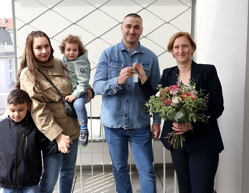 Eine Familie mit 2 kleinen Kindern steht neben einer Person, die einen Blumenstrauß in der Hand hält. Die weibliche Person der Familie hält ein Kind auf dem Arm und das zweite an der Hand, der Vater bekommt den Schlüssel von der Frau mit dem Blumenstrauß gereicht.
