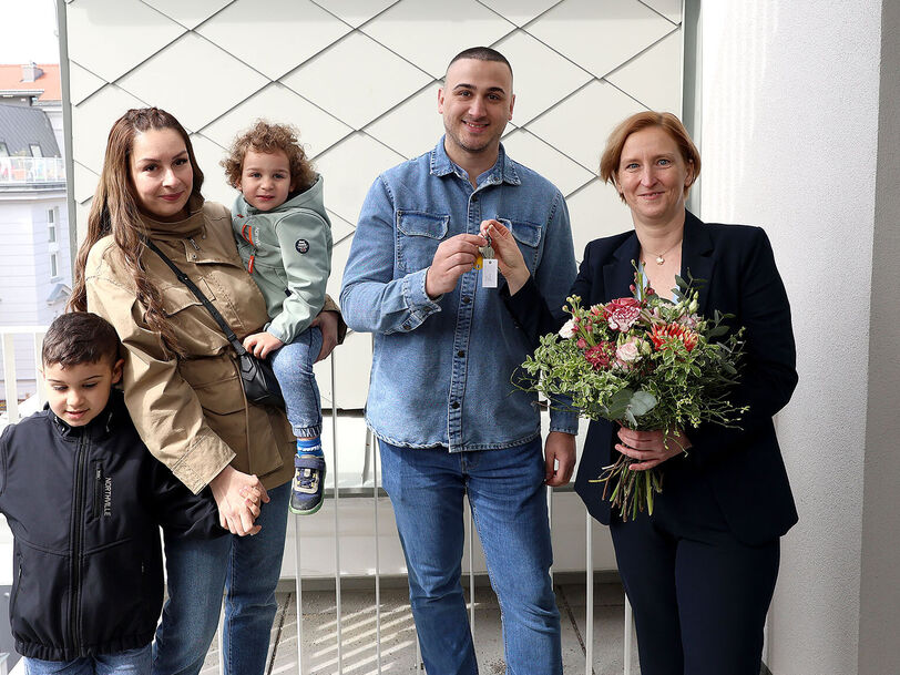 Eine Familie mit 2 kleinen Kindern steht neben einer Person, die einen Blumenstrauß in der Hand hält. Die weibliche Person der Familie hält ein Kind auf dem Arm und das zweite an der Hand, der Vater bekommt den Schlüssel von der Frau mit dem Blumenstrauß gereicht.