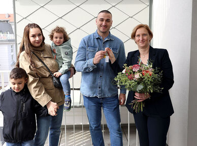 Eine Familie mit 2 kleinen Kindern steht neben einer Person, die einen Blumenstrauß in der Hand hält. Die weibliche Person der Familie hält ein Kind auf dem Arm und das zweite an der Hand, der Vater bekommt den Schlüssel von der Frau mit dem Blumenstrauß gereicht.