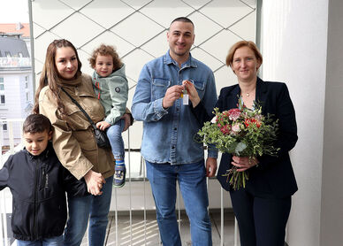 Eine Familie mit 2 kleinen Kindern steht neben einer Person, die einen Blumenstrauß in der Hand hält. Die weibliche Person der Familie hält ein Kind auf dem Arm und das zweite an der Hand, der Vater bekommt den Schlüssel von der Frau mit dem Blumenstrauß gereicht.