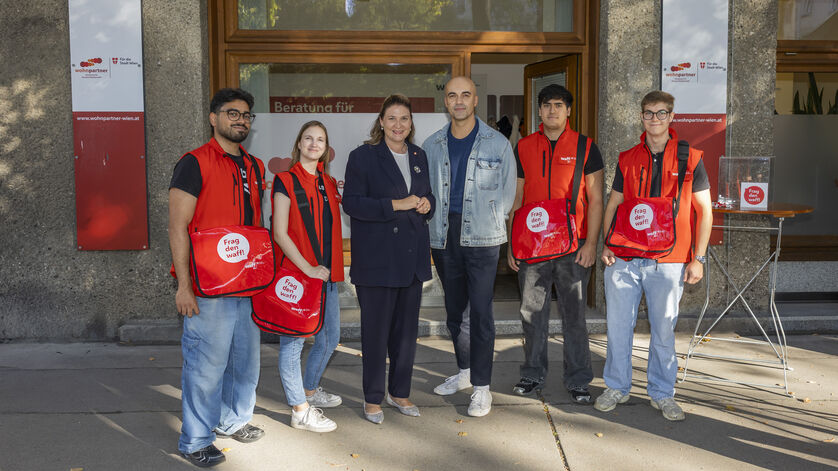 Fünf junge Menschen mit roten Westen und Taschen sowie zwei Erwachsene stehen vor einem Gebäude mit Schildern und einem Schild mit der Aufschrift wohnpartner