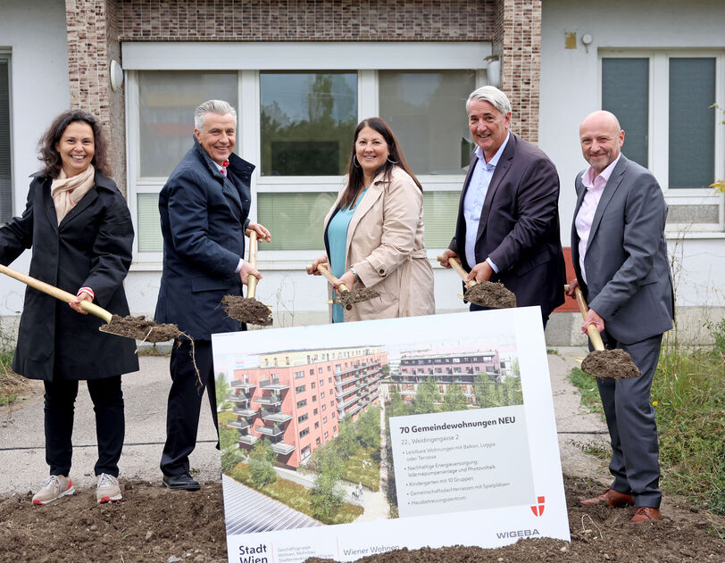 Katharina Klement (Vizedirektorin von Wiener Wohnen), Gemeinderat Josef Taucher, Vizebürgermeisterin und Wohnbaustadträtin Kathrin Gaál, Bezirksvorsteher Ernst Nevrivy und Paul Steurer, Direktor der WIGEBA, stehen jeweils mit einem Spaten voll Erde hinter einer Tafel mit der Visualisierung des Gemeindebau NEU in der Weidingergasse. Sie sind durchwegs herbstlich gekleidet und lächeln in die Kamera. 