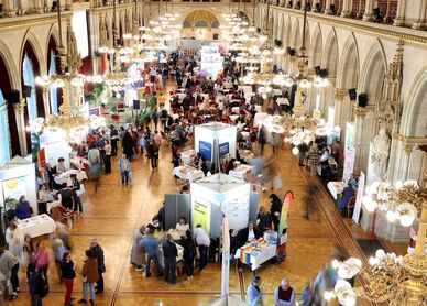 Der große Festsaal im Wiener Rathaus, aufgenommen von einer erhobenen Position. Der langgezogene Saal ist im Neogotischen Stil errichtet und mit prächtigen goldenen Lustern ausgestattet. Zu erkennen sind Messestände auf dem Parkettboden und viele Besucher*innen. 