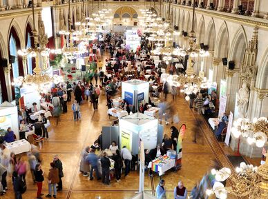 Der große Festsaal im Wiener Rathaus, aufgenommen von einer erhobenen Position. Der langgezogene Saal ist im Neogotischen Stil errichtet und mit prächtigen goldenen Lustern ausgestattet. Zu erkennen sind Messestände auf dem Parkettboden und viele Besucher*innen. 