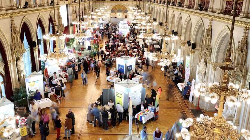 Der große Festsaal im Wiener Rathaus, aufgenommen von einer erhobenen Position. Der langgezogene Saal ist im Neogotischen Stil errichtet und mit prächtigen goldenen Lustern ausgestattet. Zu erkennen sind Messestände auf dem Parkettboden und viele Besucher*innen. 
