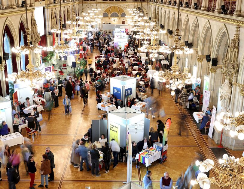 Der große Festsaal im Wiener Rathaus, aufgenommen von einer erhobenen Position. Der langgezogene Saal ist im Neogotischen Stil errichtet und mit prächtigen goldenen Lustern ausgestattet. Zu erkennen sind Messestände auf dem Parkettboden und viele Besucher*innen. 