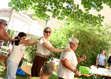 Mehrere Menschen tanzen in einer Reihe im Freien unter Bäumen bei einem Fest mit Tischen und Zelten im Hintergrund