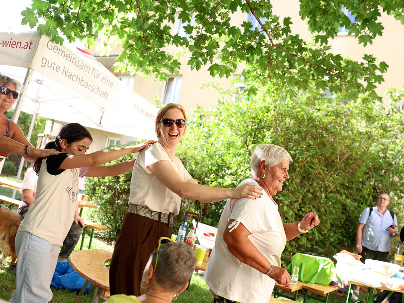 Mehrere Menschen tanzen in einer Reihe im Freien unter Bäumen bei einem Fest mit Tischen und Zelten im Hintergrund