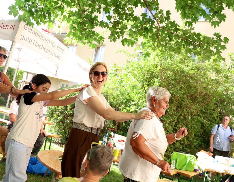 Mehrere Menschen tanzen in einer Reihe im Freien unter Bäumen bei einem Fest mit Tischen und Zelten im Hintergrund