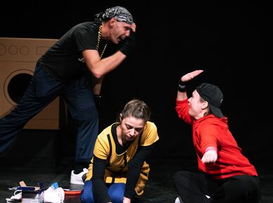 Szenenbild aus dem Gemeindebautheater: Ein schwarz gekleideter Jugendlicher mit Kopftuch gibt einer auf dem Boden sitzenden Jugendlichen mit rotem Sweater und verkehrt getragenem schwarzen Baseballcap High Five. Dazwischen kniet eine Jugendlich mit gelbem Oberteil und zeichnet mit Kreide etwas auf den Boden.