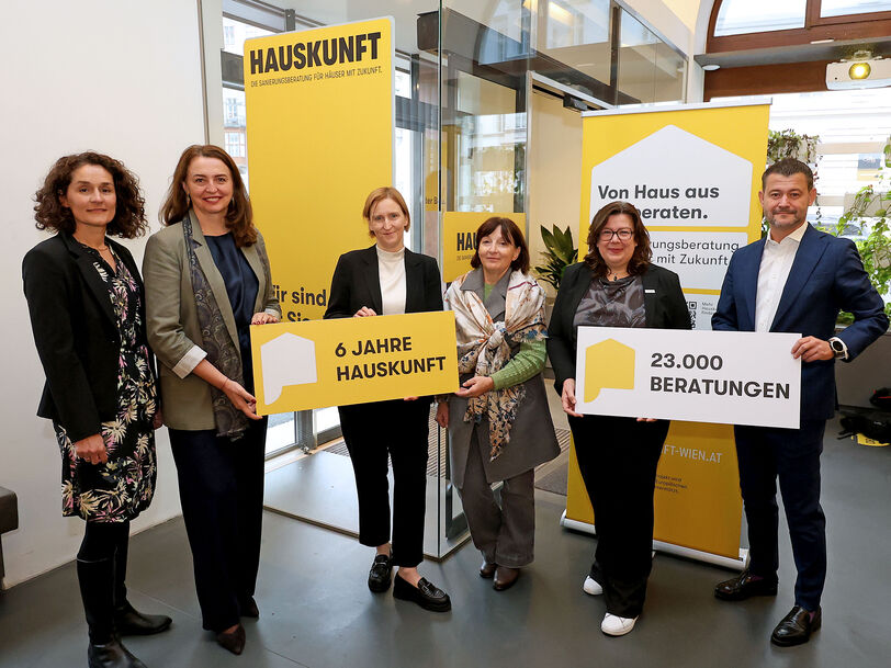 Fünf Personen stehen in einem modernen Innenraum und halten gelbe Schilder mit den Aufschriften '6 JAHRE HAUSKUNFT' und '23.000 BERATUNGEN', im Hintergrund gelbe Banner mit dem Schriftzug 'HAUSKUNFT'