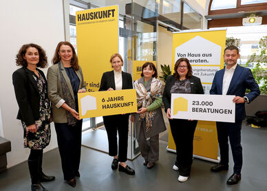 Fünf Personen stehen in einem modernen Innenraum und halten gelbe Schilder mit den Aufschriften '6 JAHRE HAUSKUNFT' und '23.000 BERATUNGEN', im Hintergrund gelbe Banner mit dem Schriftzug 'HAUSKUNFT'