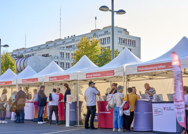 Menschen stehen an mehreren weißen Zelten mit roten Bannern und Informationsständen in einer sonnigen Straße vor einem großen Gebäude