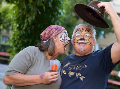 Eine Frau und ein Mann sind wie ein Hund und ein Tiger geschminkt, der Hund (die Frau) bellt offenbar den Tiger an, der freundlich in die Kamera lächelt und dabei seinen Hut zieht.