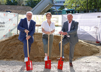 Drei Personen in formeller Kleidung stehen auf einer Baustelle und halten jeweils einen roten Spaten in der Hand, hinter ihnen ein großer Erdhaufen und Bauzäune mit dem Logo der Stadt Wien