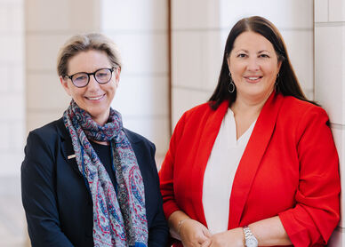 Zwei Frauen, Karin Ramser und Kathrin Gaál, stehen in einem Gang des Rathauses und lächeln in die Kamera. Beide sind businessmäßig mit Blazern gekleidet.