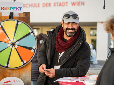 Ein freundlich lächelnder Mitarbeiter von wohnpartner, erkennbar an seinem Patch, steht neben einem Glücksrad . Er ist offensichtlich für kühleres Wetter gekleidet.