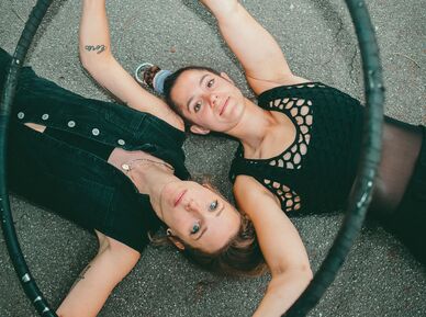Zwei junge Frauen liegen Kopf neben Kopf in gegengesetzter Richtung auf dem Asphalt und blicken in die Kamera. Sie sind mit schwarzen Shirts und schwarzen Leggings bekleidet und halten einen Hulahoop-Ring in ihren Händen.