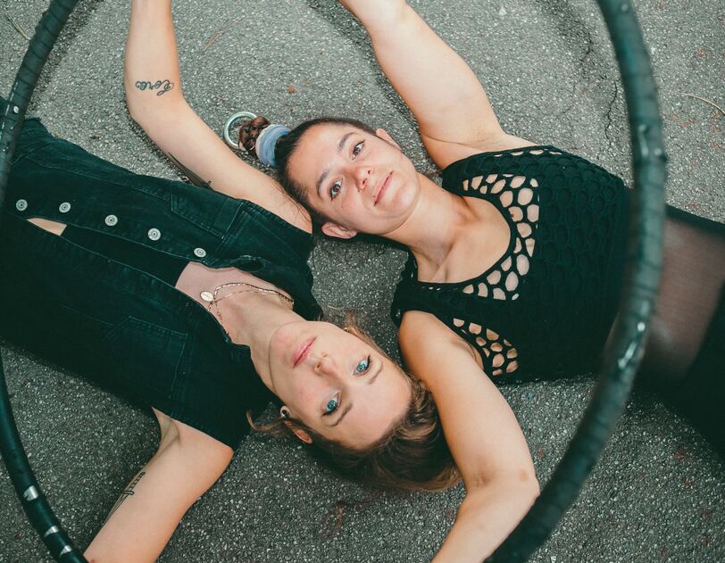 Zwei junge Frauen liegen Kopf neben Kopf in gegengesetzter Richtung auf dem Asphalt und blicken in die Kamera. Sie sind mit schwarzen Shirts und schwarzen Leggings bekleidet und halten einen Hulahoop-Ring in ihren Händen.