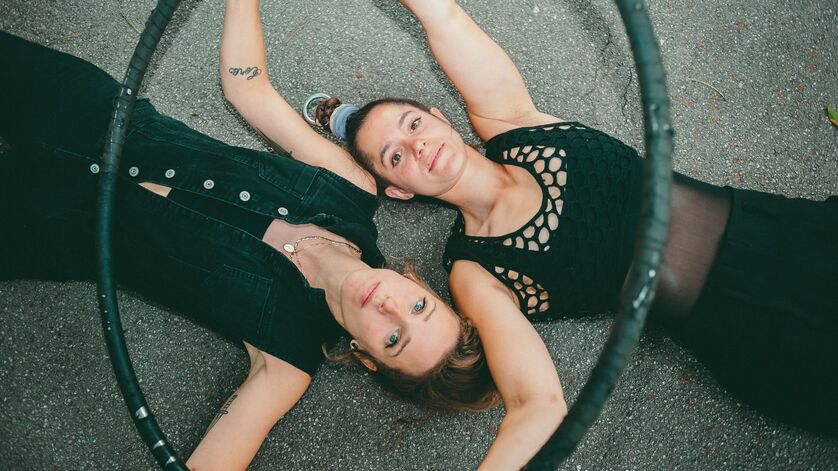Zwei junge Frauen liegen Kopf neben Kopf in gegengesetzter Richtung auf dem Asphalt und blicken in die Kamera. Sie sind mit schwarzen Shirts und schwarzen Leggings bekleidet und halten einen Hulahoop-Ring in ihren Händen.