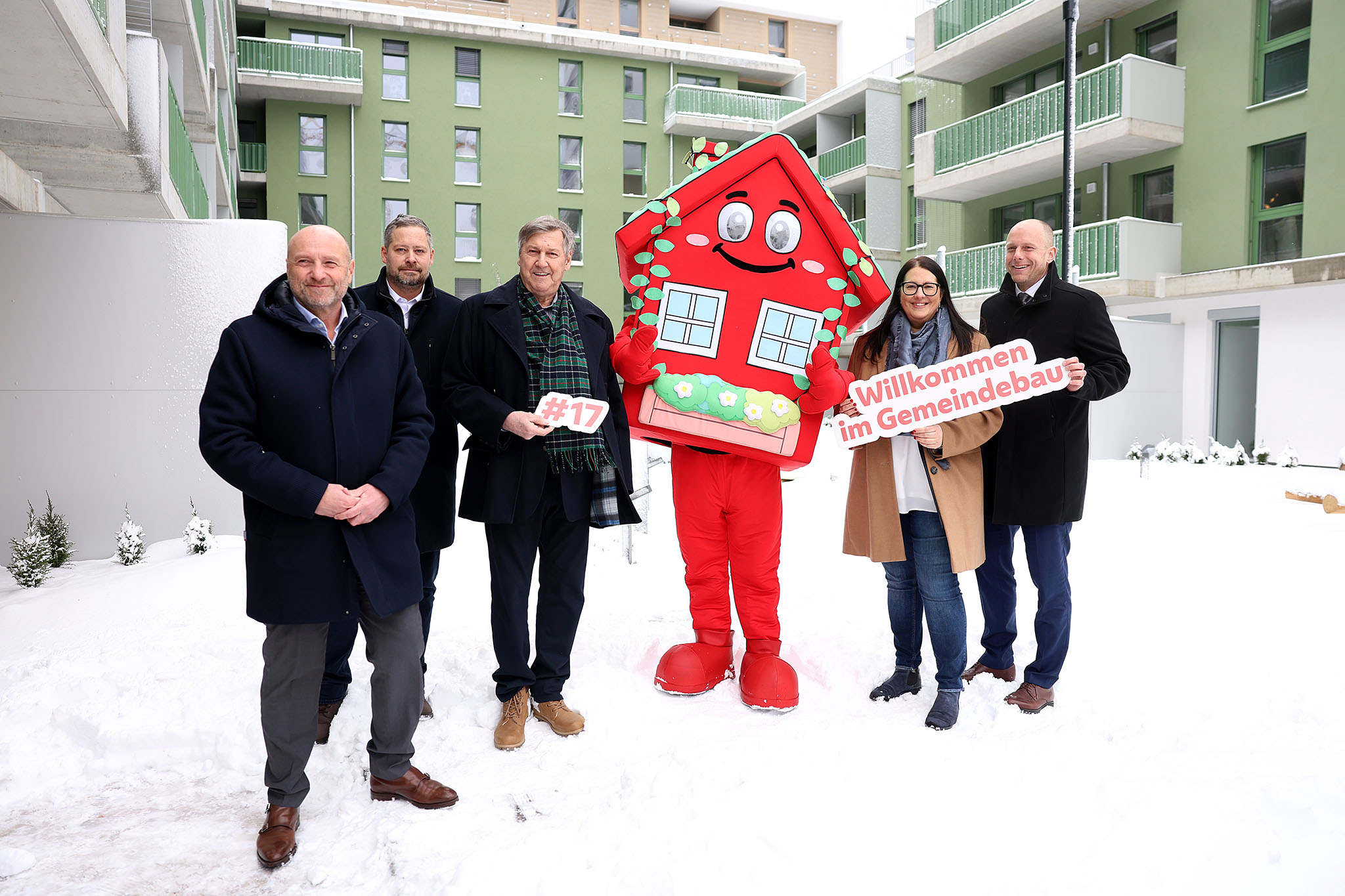 5 Personen stehen in winterlicher Business-Bekleidung im verschneiten Innenhof einer Neubauanlage. Ihnen zur Seite wurde wurde Wohna Lisa, das Maskottchen des geförderten Wohnbaus in Wien. Es ist eine als Haus verleidete rote Figur in Form eines Hauses mit Satteldach, großen Augen und einem freundlichen Lächeln.